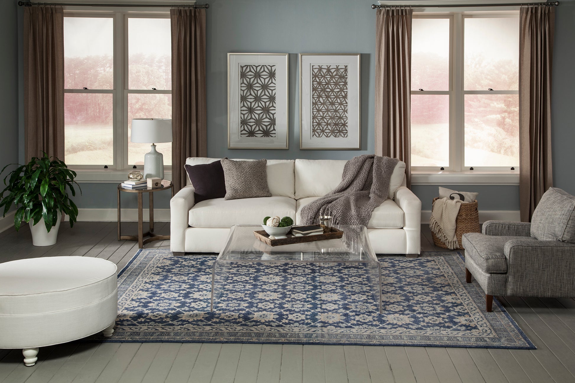 Modern living room with white sofa, gray armchair, clear acrylic coffee table, blue patterned rug, brown curtains, plant.