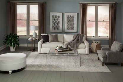 Neutral living room with white sofa, gray armchair, clear acrylic coffee table, light gray rug, two windows with curtains.