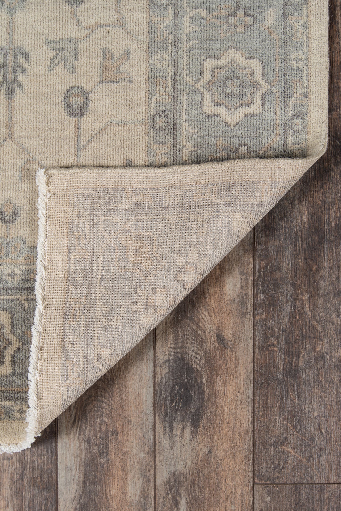 Beige and blue oriental rug with faded medallion pattern; folded corner shows underside on rustic wooden floor.