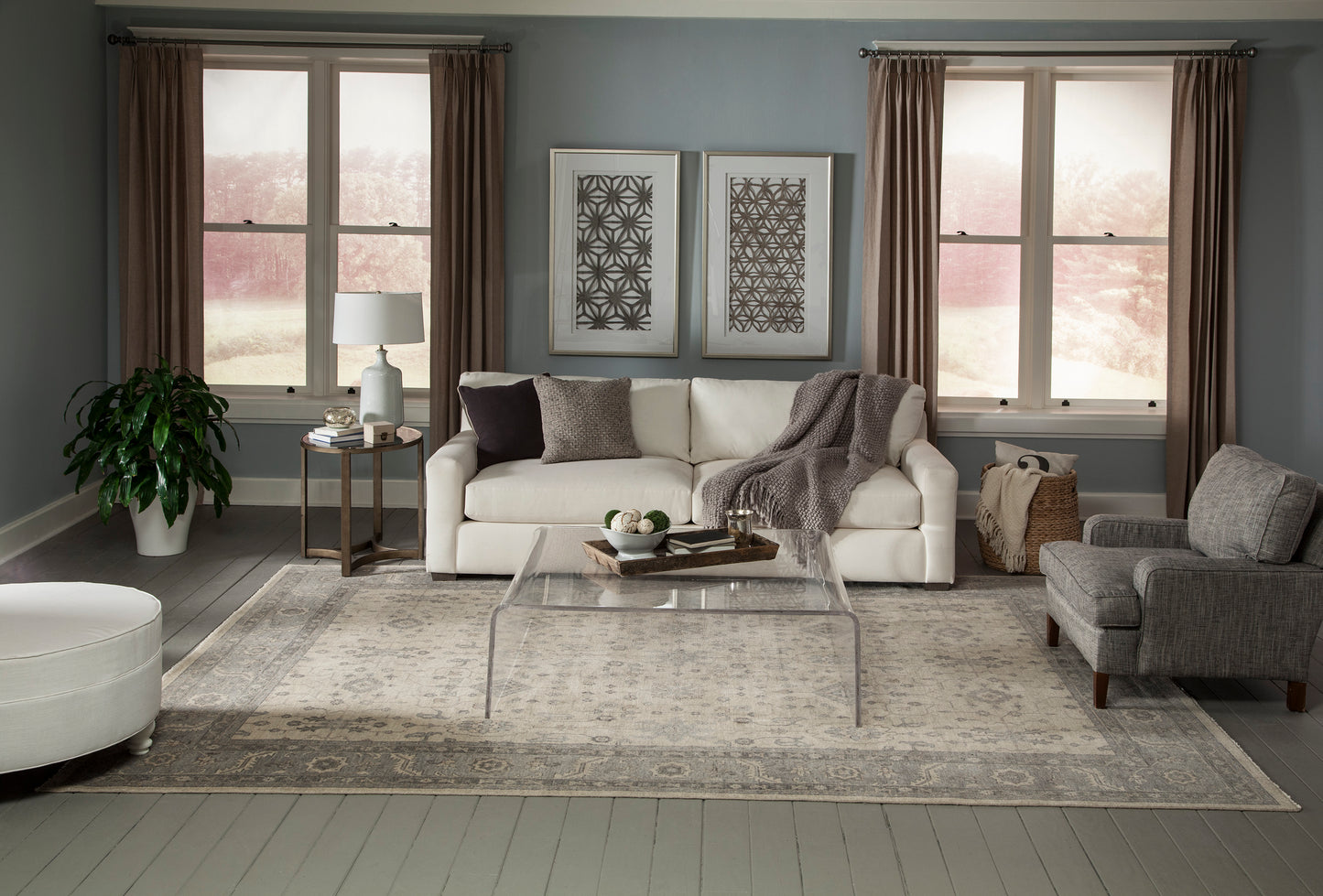 White sofa, gray armchair, clear glass coffee table, beige rug, potted plant, two framed prints, and brown drapes.