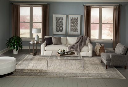 White sofa, gray armchair, clear glass coffee table, beige rug, potted plant, two framed prints, and brown drapes.
