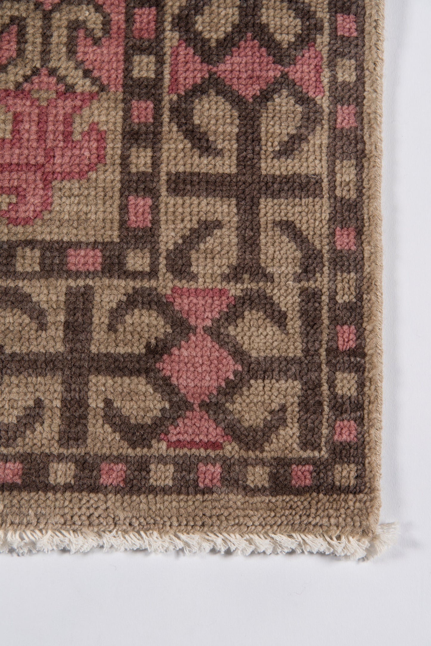 Geometric woven rug with tan background, dark brown diamond patterns and pink accents; wool texture, fringed edge.