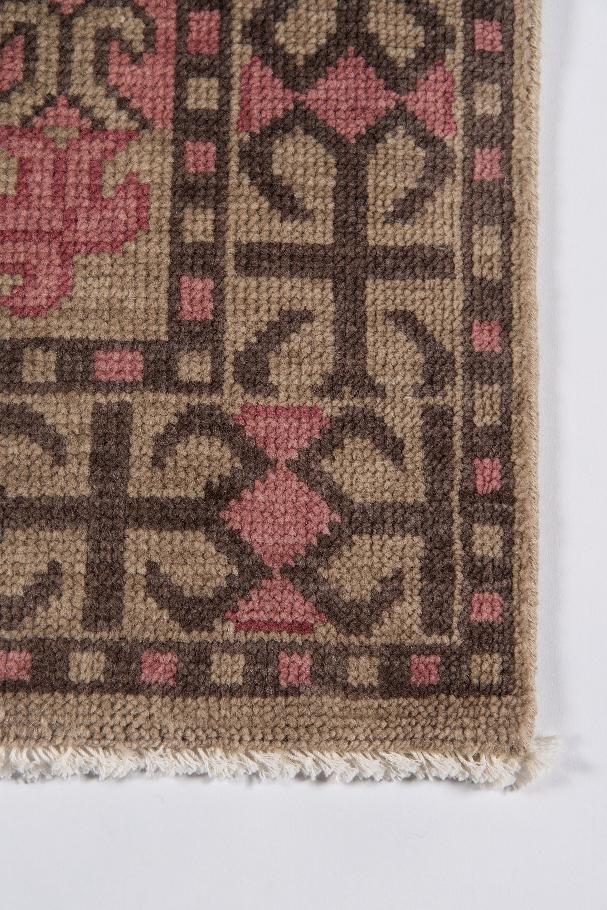 Geometric woven rug with tan background, dark brown diamond patterns and pink accents; wool texture, fringed edge.