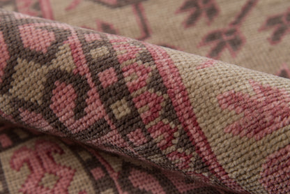 Close-up of a beige woven rug with pink and brown geometric motifs, textured pile, rustic kilim-style decor.