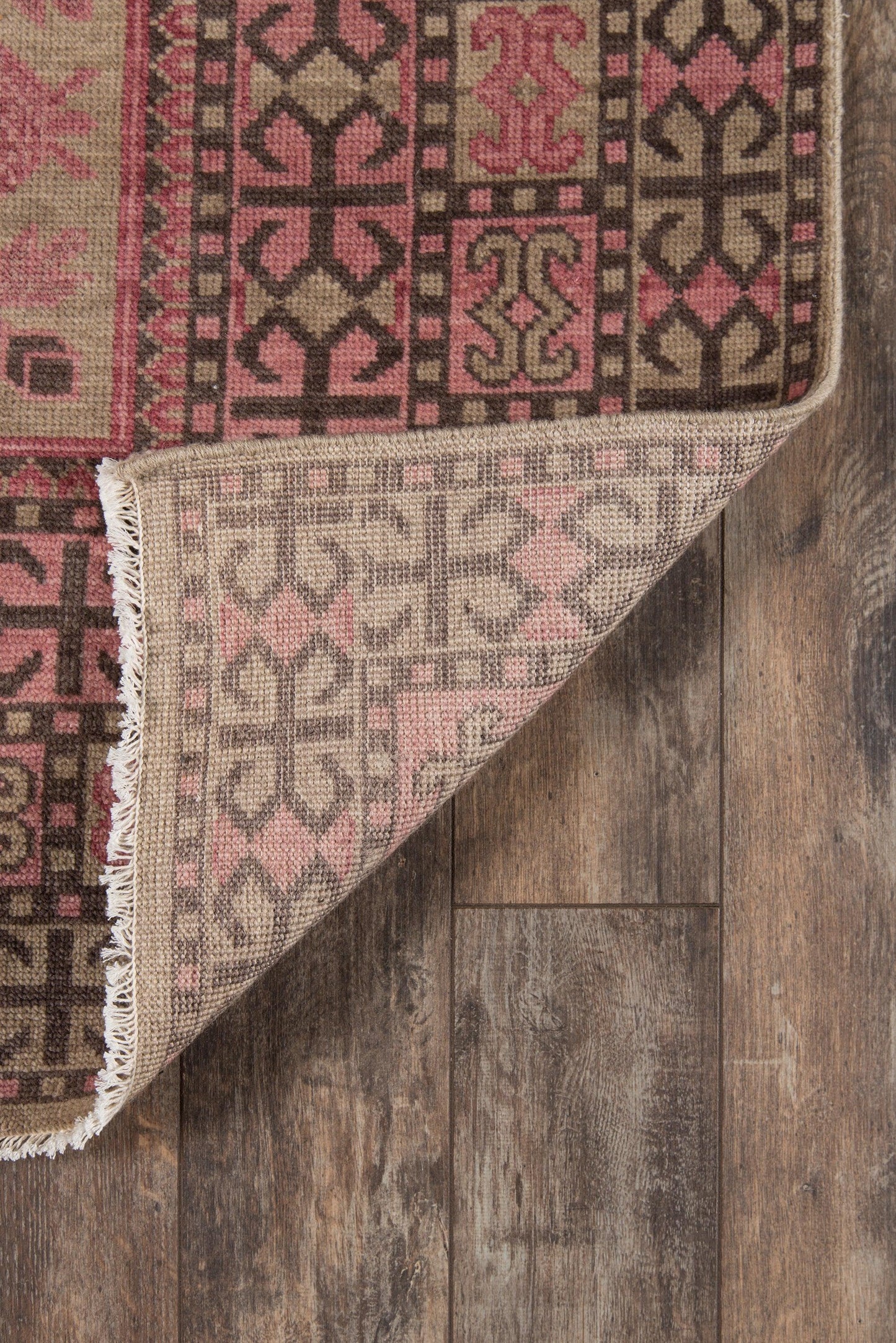 Woven geometric rug in pink, brown and beige with fringe, shown on dark wood floor.