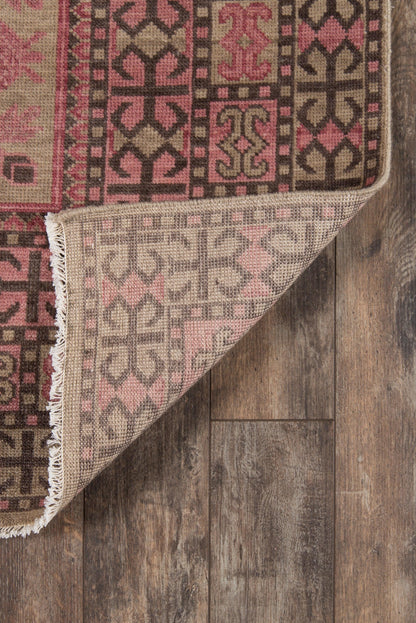 Woven geometric rug in pink, brown and beige with fringe, shown on dark wood floor.