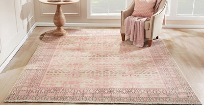 Soft pink traditional area rug with beige base and intricate borders, in a sunny living room beside a beige armchair.