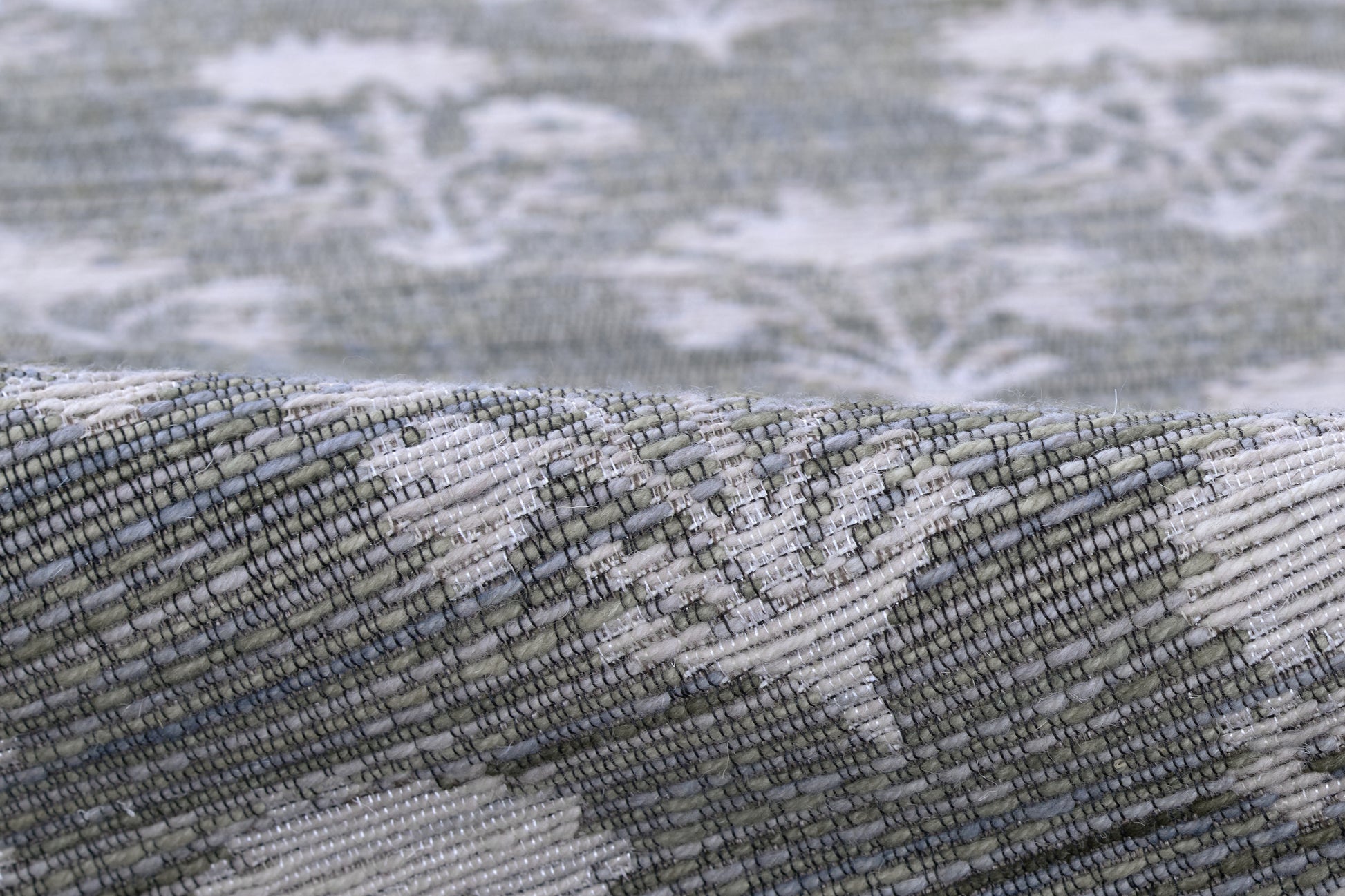 Close-up of a gray-blue woven rug with white damask pattern, textured weave, and folded edge.