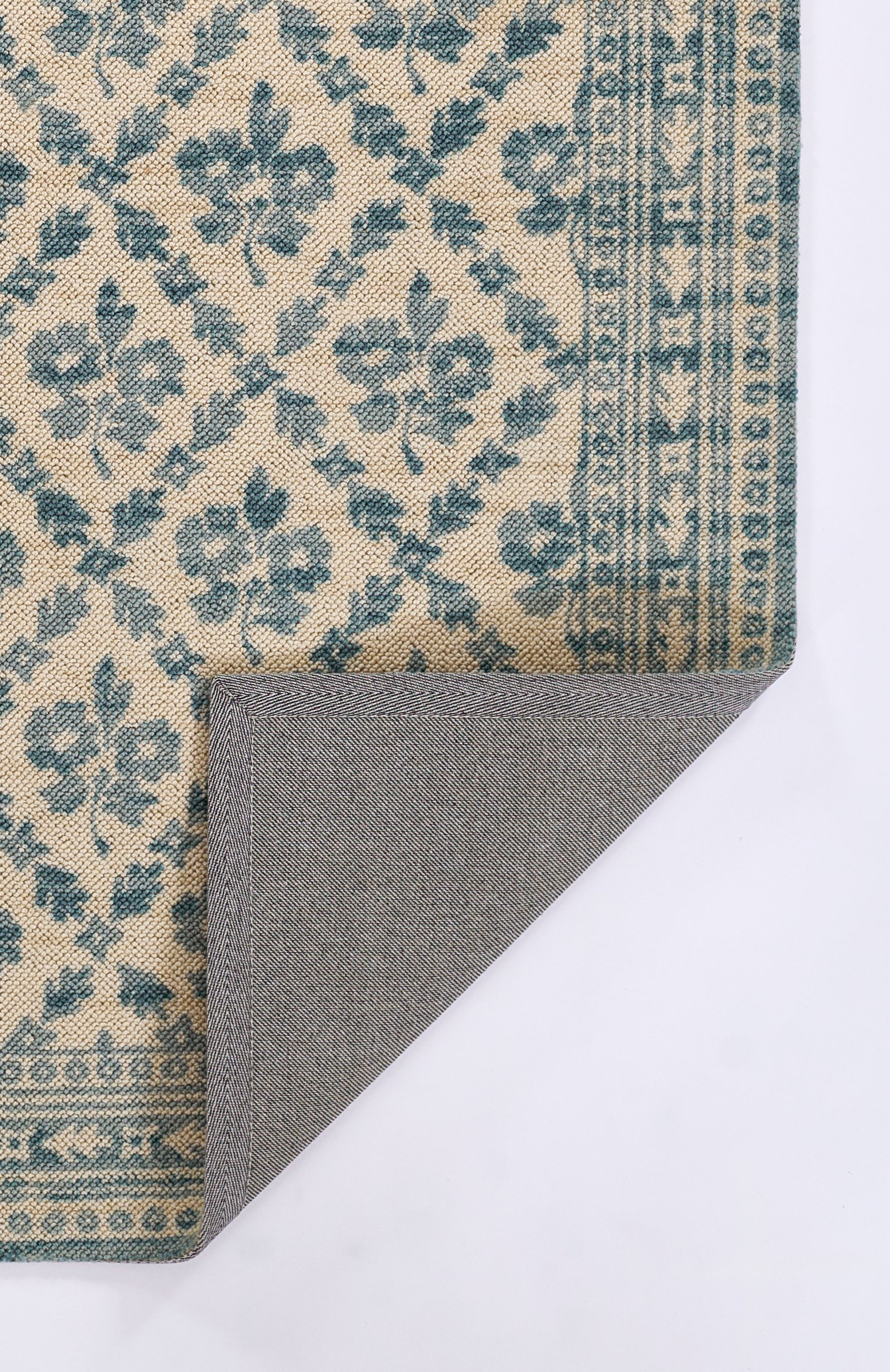 Beige rug with blue floral pattern and border; folded corner reveals grey herringbone backing.