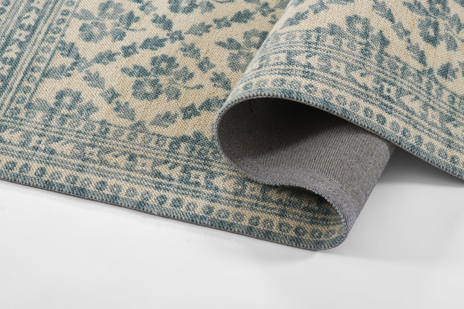 Teal and beige patterned area rug with floral border, folded corner showing grey backing.