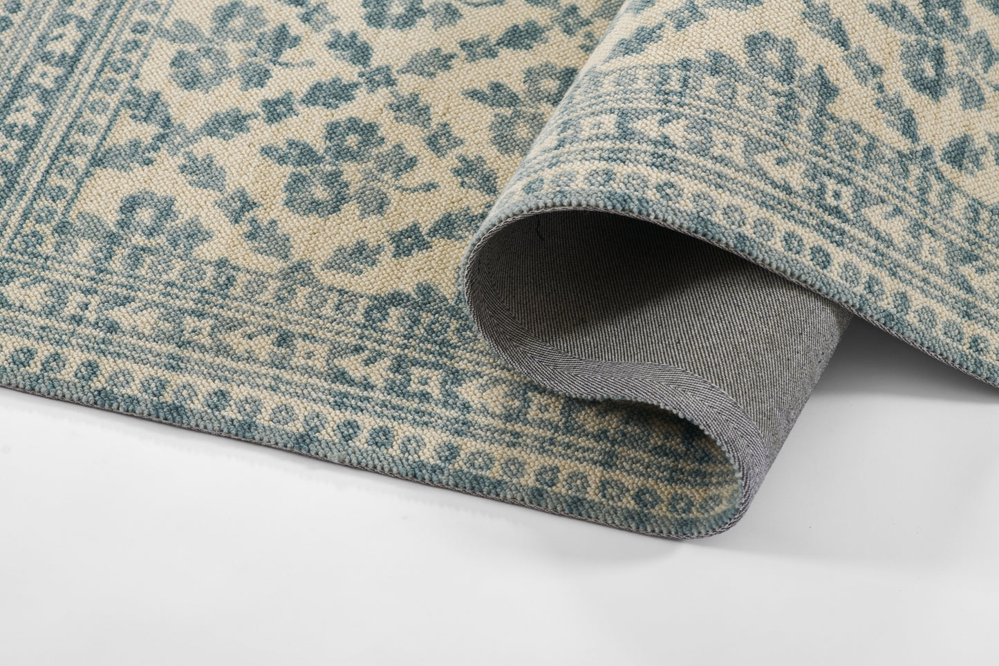 Blue-teal and beige floral rug with woven texture; rolled edge reveals gray backing.