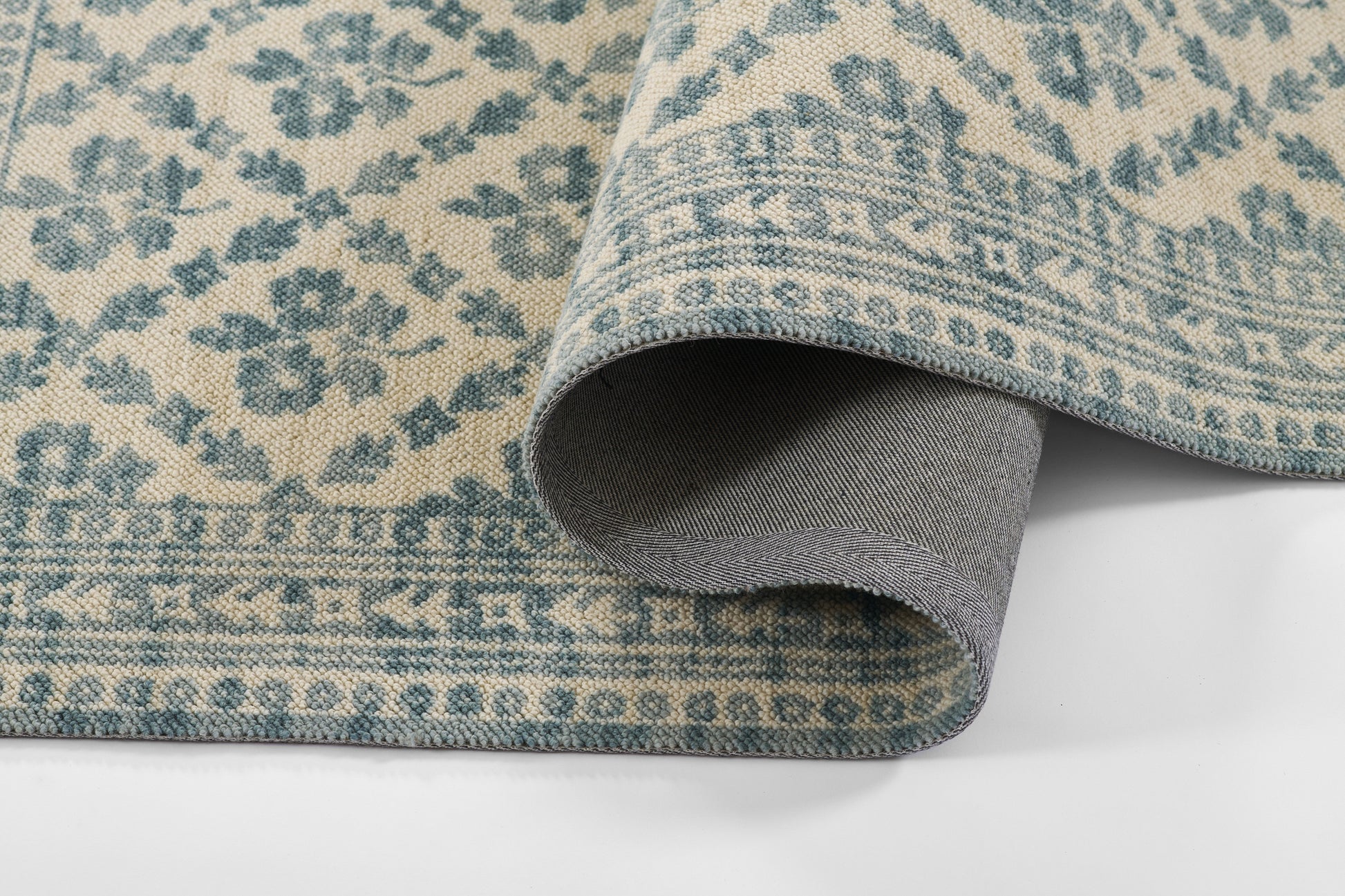 Blue and beige traditional floral rug with ornate border, rolled to show grey backing.