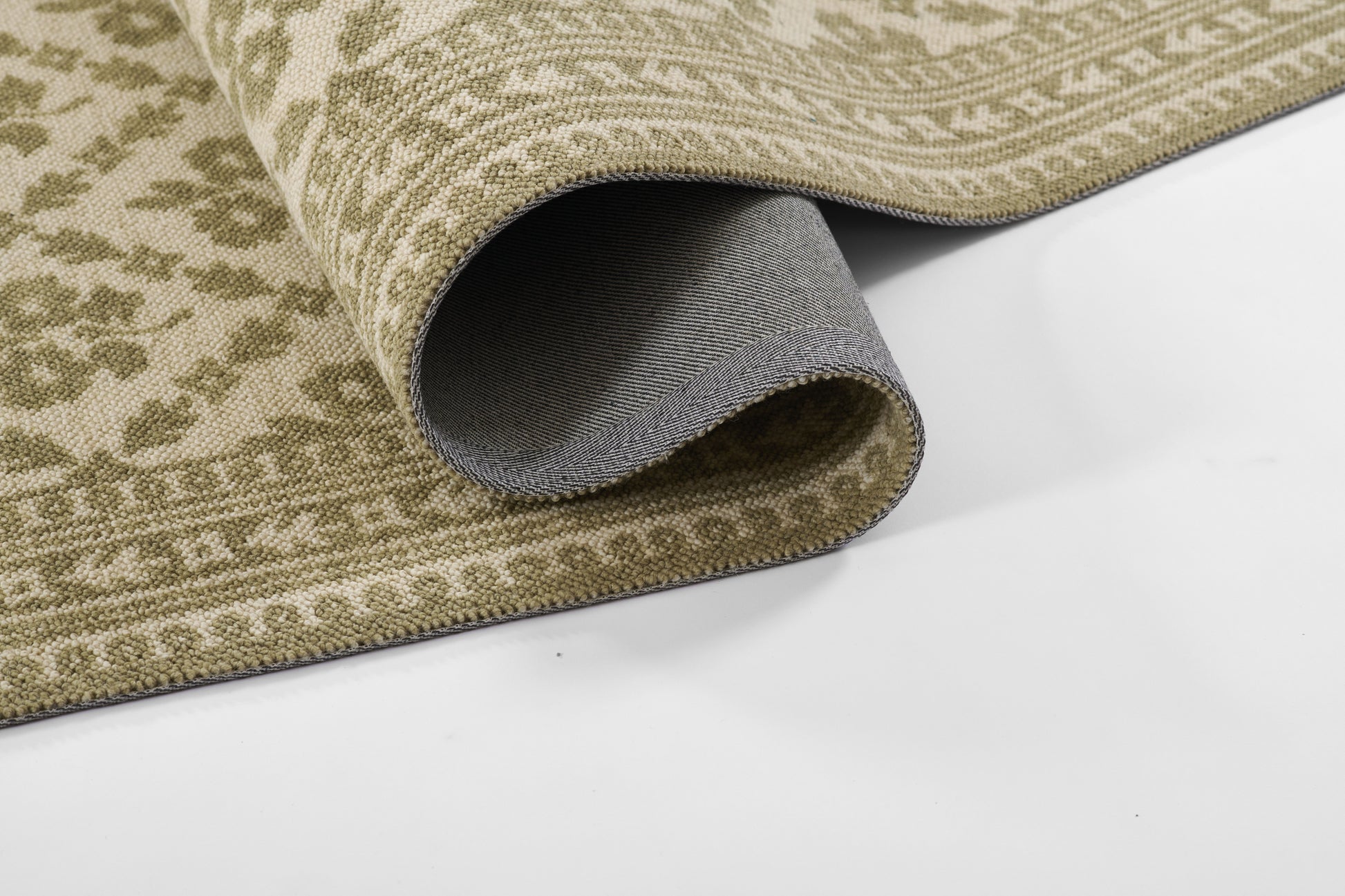 Beige and sage-green patterned area rug, rolled edge reveals gray underside; tight woven texture.