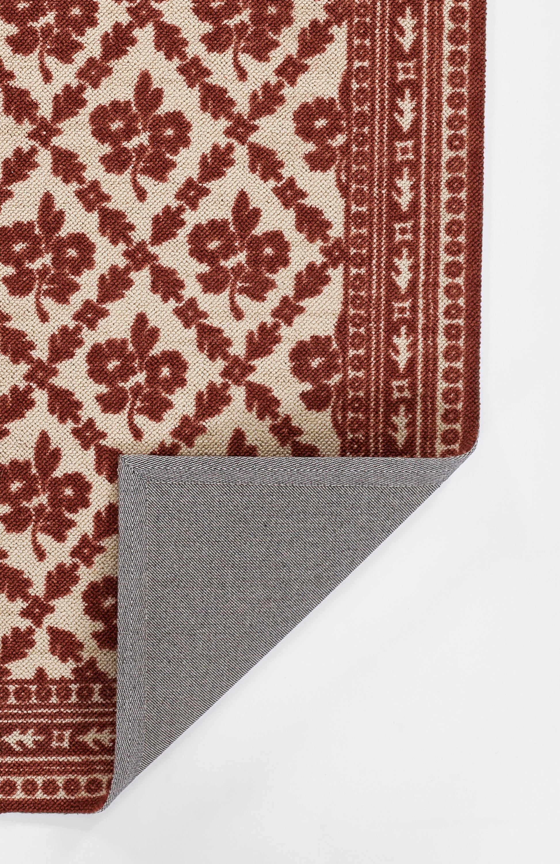 Red and cream floral patterned rug with a decorative border; grey herringbone reverse peeks at corner.