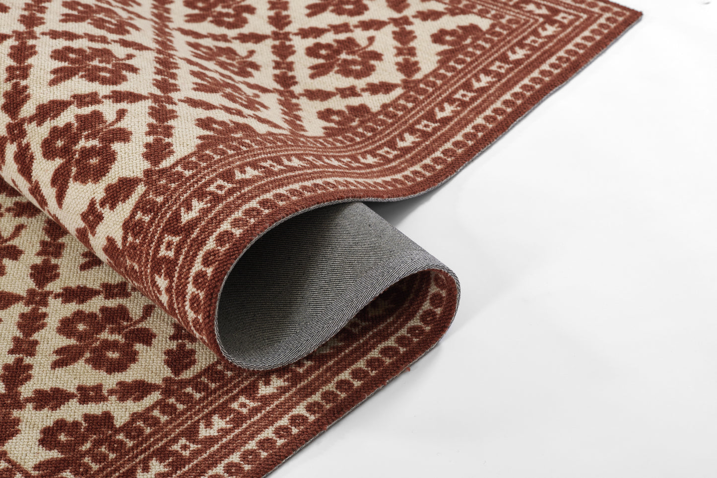 Beige rug with deep red ornate floral pattern and border; rolled corner reveals grey backing.