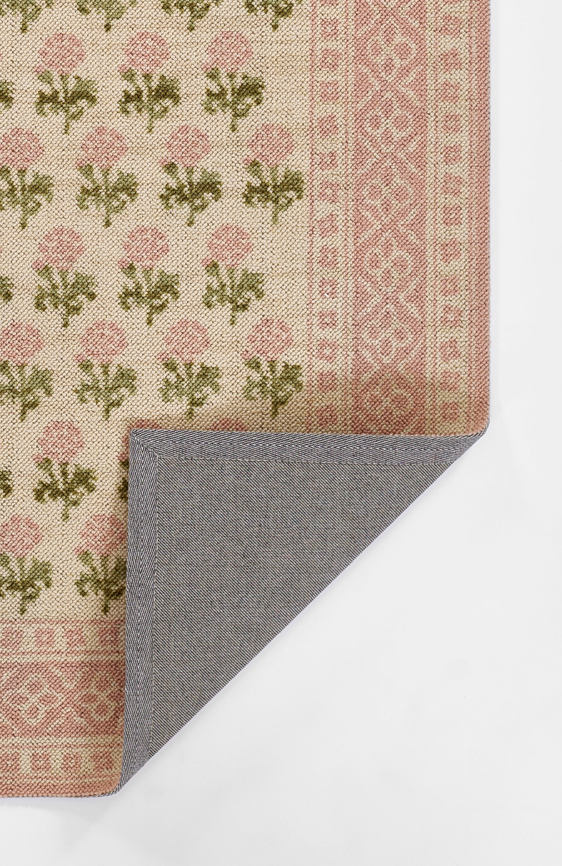 Beige rug with pink floral motifs and border, green leaves; folded corner reveals blue-gray herringbone reverse.