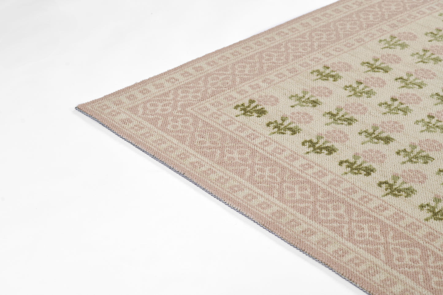 Rectangular pink-beige woven rug with green floral motifs and diamond geometric border; fringe edge visible.