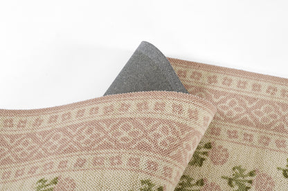 Soft pink and cream geometric woven rug with subtle green accents; folded corner shows blue denim-like backing.