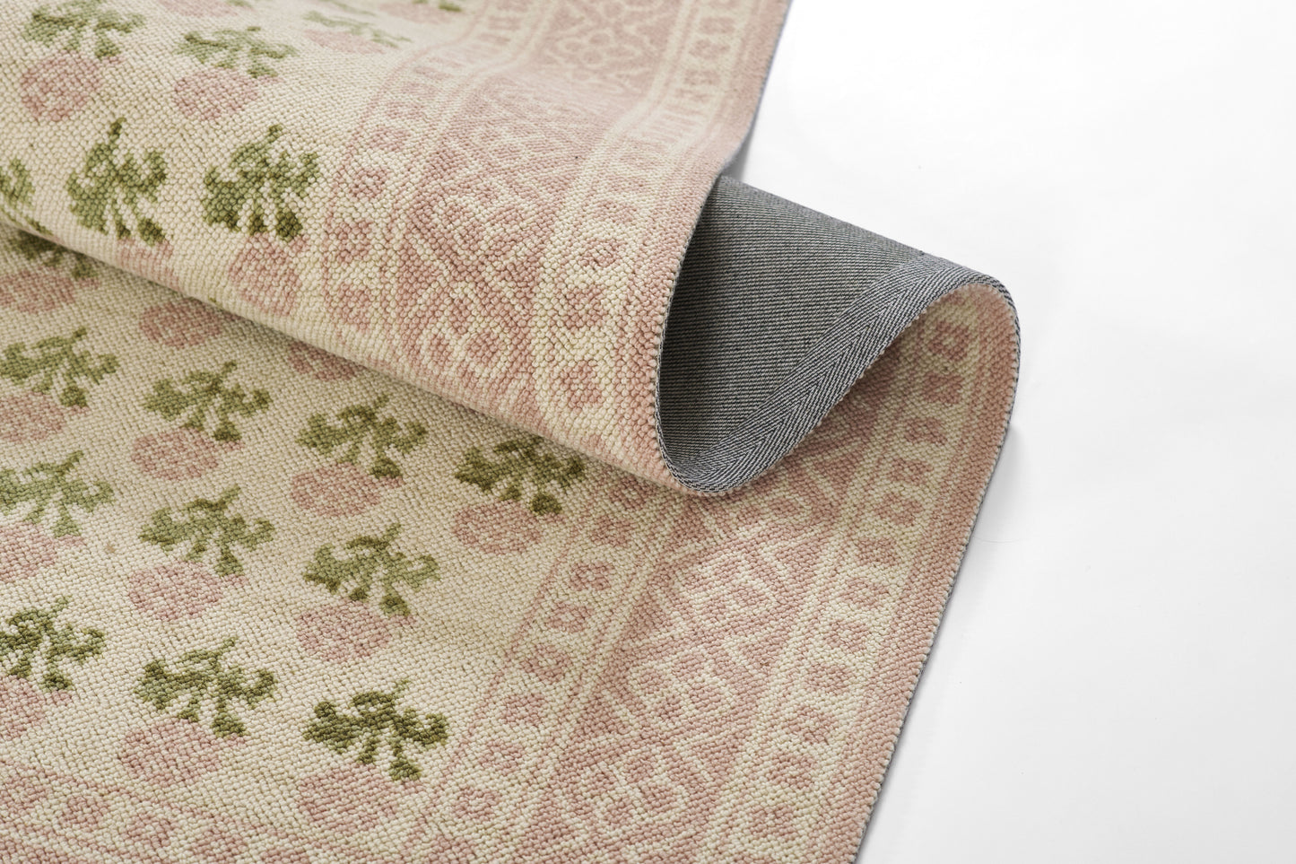 Beige woven rug with pink floral pattern and green leaves; pink geometric border, partially rolled to show gray backing