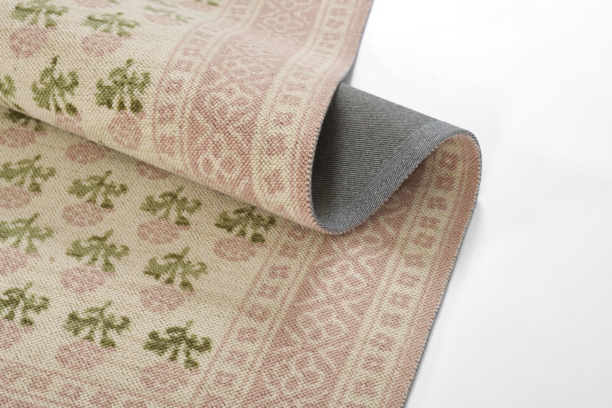 Beige woven rug with pink floral pattern and green leaves; pink geometric border, partially rolled to show gray backing