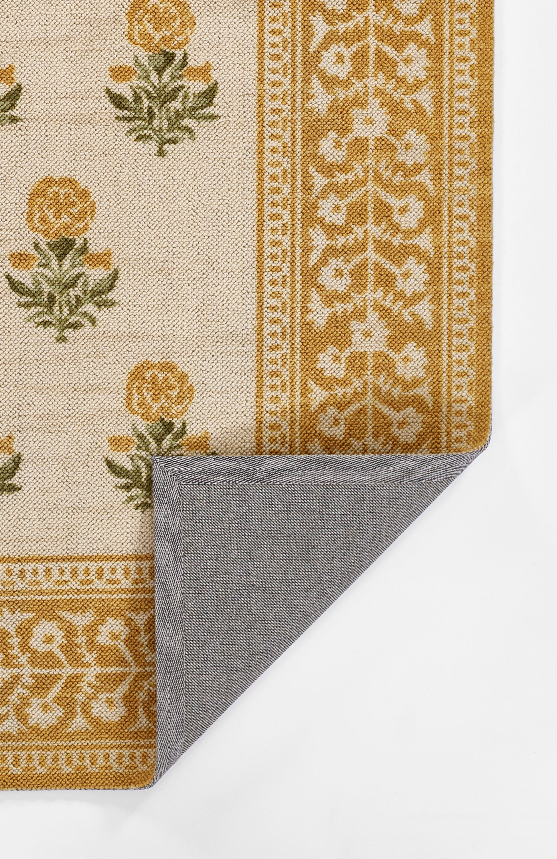 Cream-beige woven rug with mustard floral motifs and a golden border; folded corner reveals gray herringbone backing.