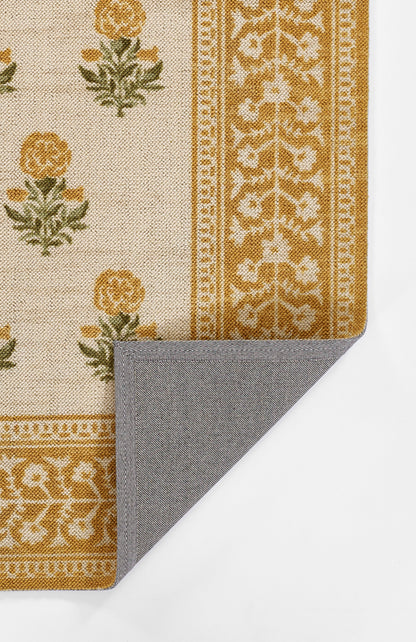 Beige rug with orange floral motifs and gold ornate border; folded corner shows grey herringbone reverse.