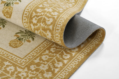 Beige woven rug in gold ornamental border with floral/pineapple motifs; partially rolled to reveal grey backing.