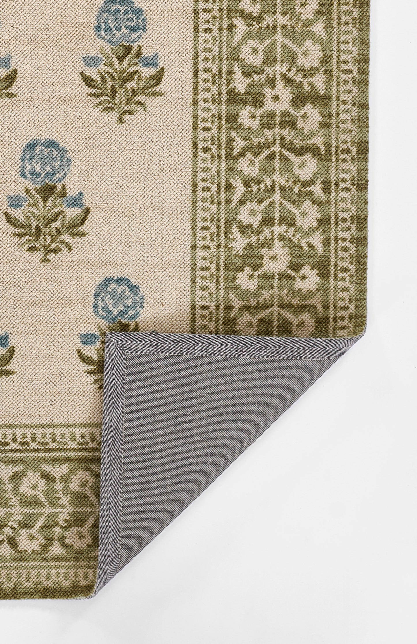 Beige floral area rug with blue blossoms and olive border; folded corner shows gray herringbone backside.