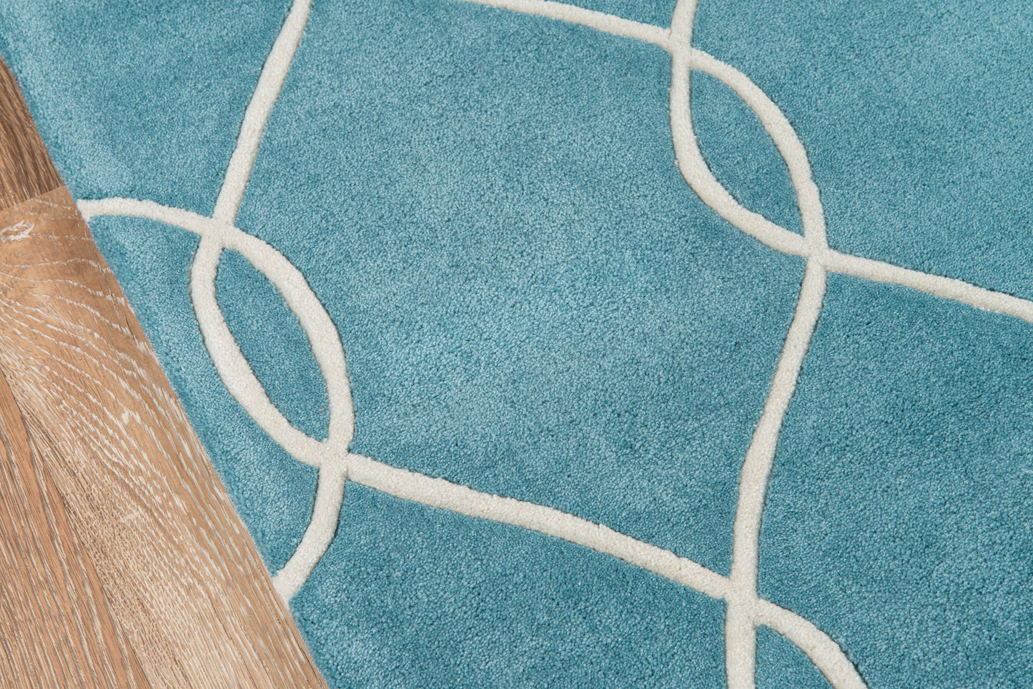 Teal rug with cream looping lines in a geometric pattern beside light wood flooring and a wooden furniture leg.