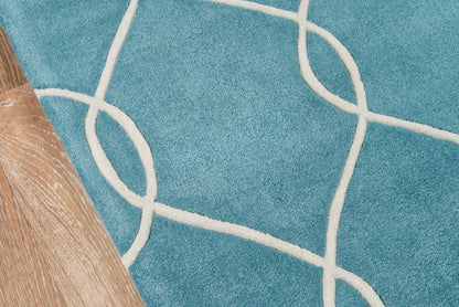 Teal rug with cream looping lines in a geometric pattern beside light wood flooring and a wooden furniture leg.