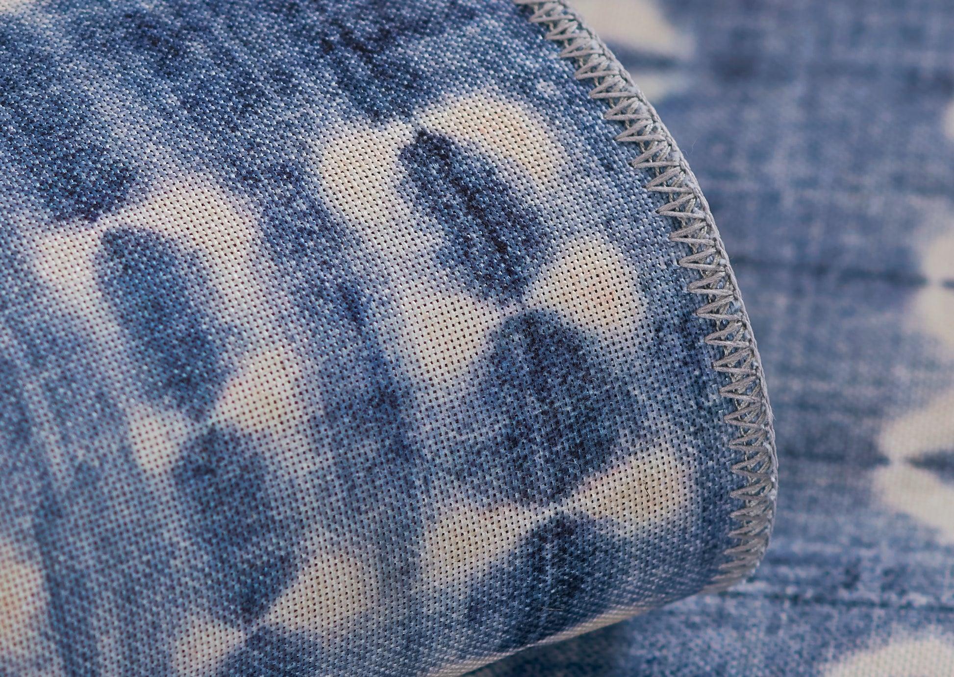 Blue and white tie-dye denim-style rug with gray overlock stitching along a curved edge.