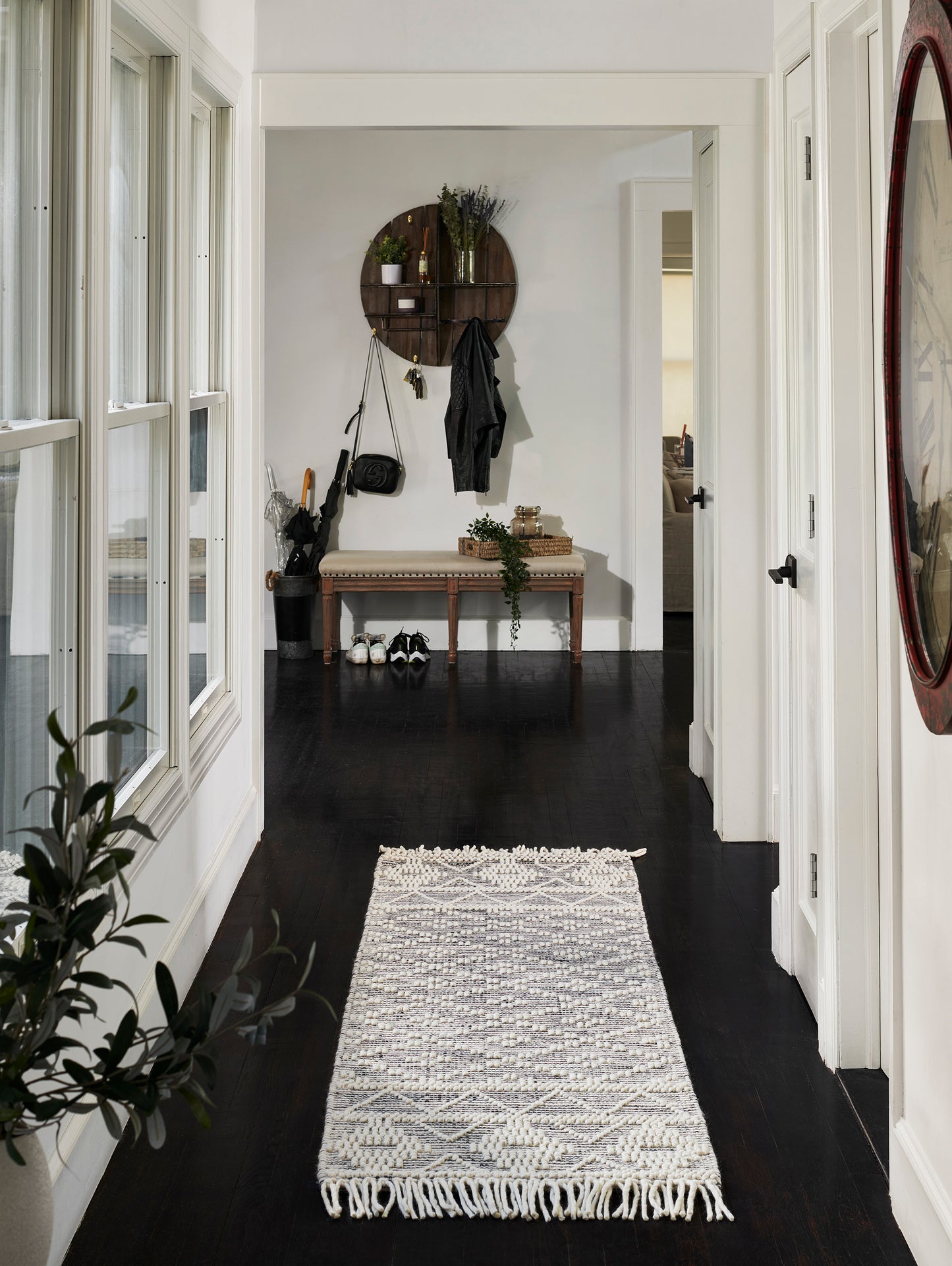 Narrow entry hall with dark hardwood floors, long woven gray patterned rug with fringe, white walls, round wall shelf, coat hooks and bench.