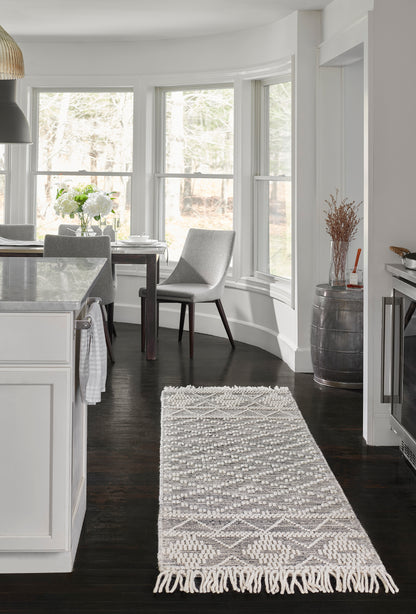 Geometric gray-and-cream woven area rug with fringe ends, placed in a bright white kitchen with dark hardwood floors.