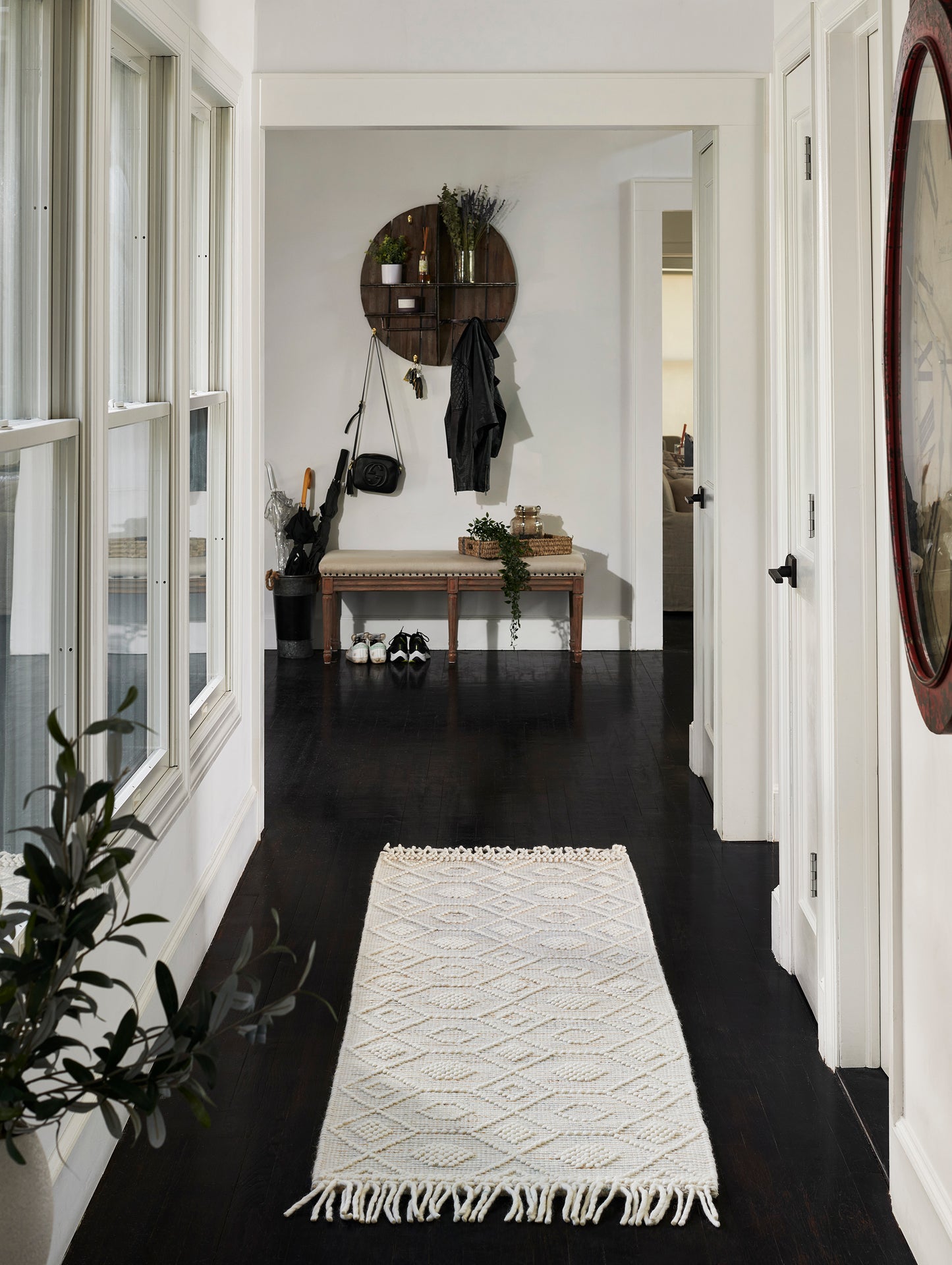 Cream diamond-patterned runner rug with fringe in a dark hardwood entry hall, beside a wooden bench and circular wall shelf.