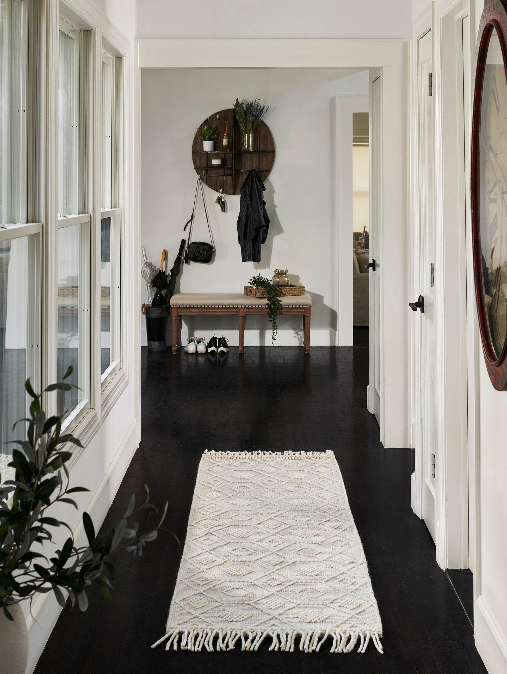 Cream woven hallway runner with fringe on dark wood floor; beige cushioned bench under a round wooden wall shelf with coats.