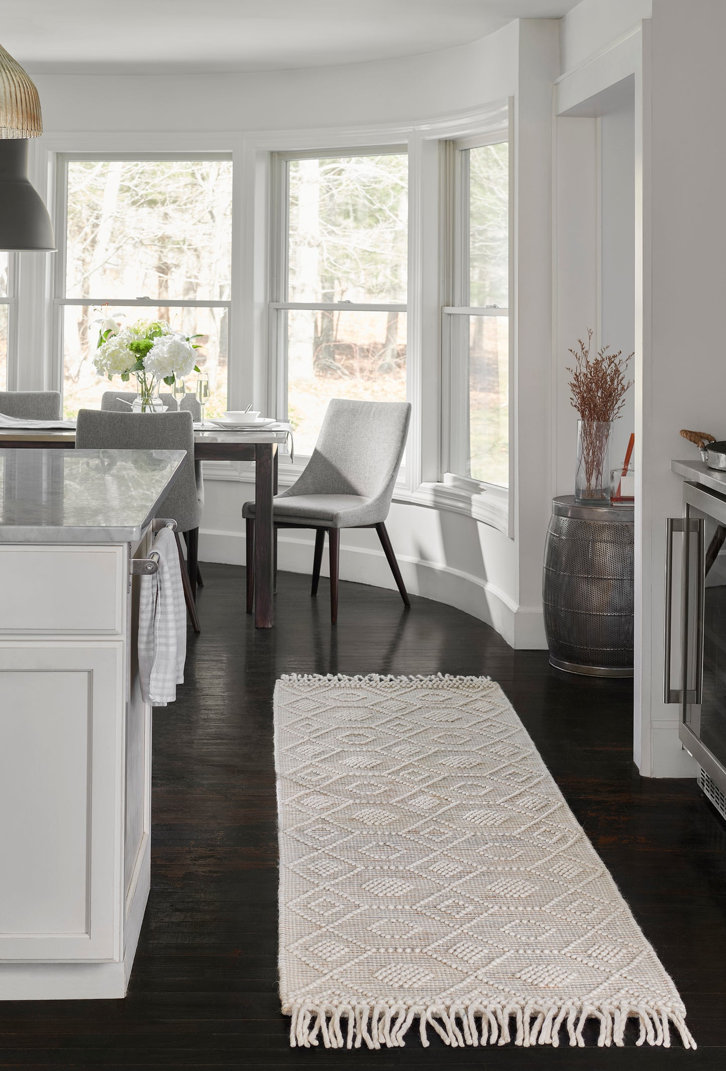 Cream woven runner rug with diamond pattern & fringed ends on dark hardwood kitchen floor by bay window dining area.