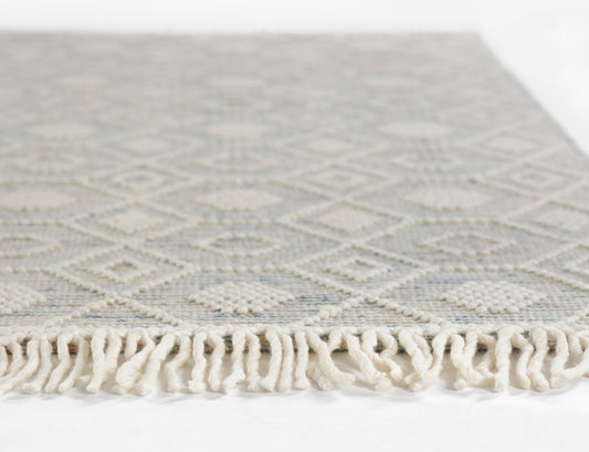 Woven rug with light blue-gray base, white diamond pattern and cream tassel fringe along edge.