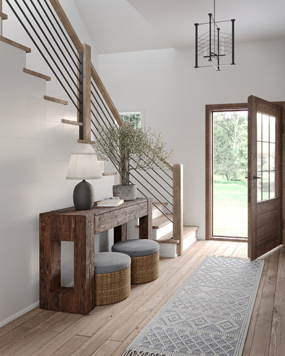 Rustic entryway with a rough wood console, two woven ottomans, a lamp, plant, staircase, glass door, and a pale patterned area rug.