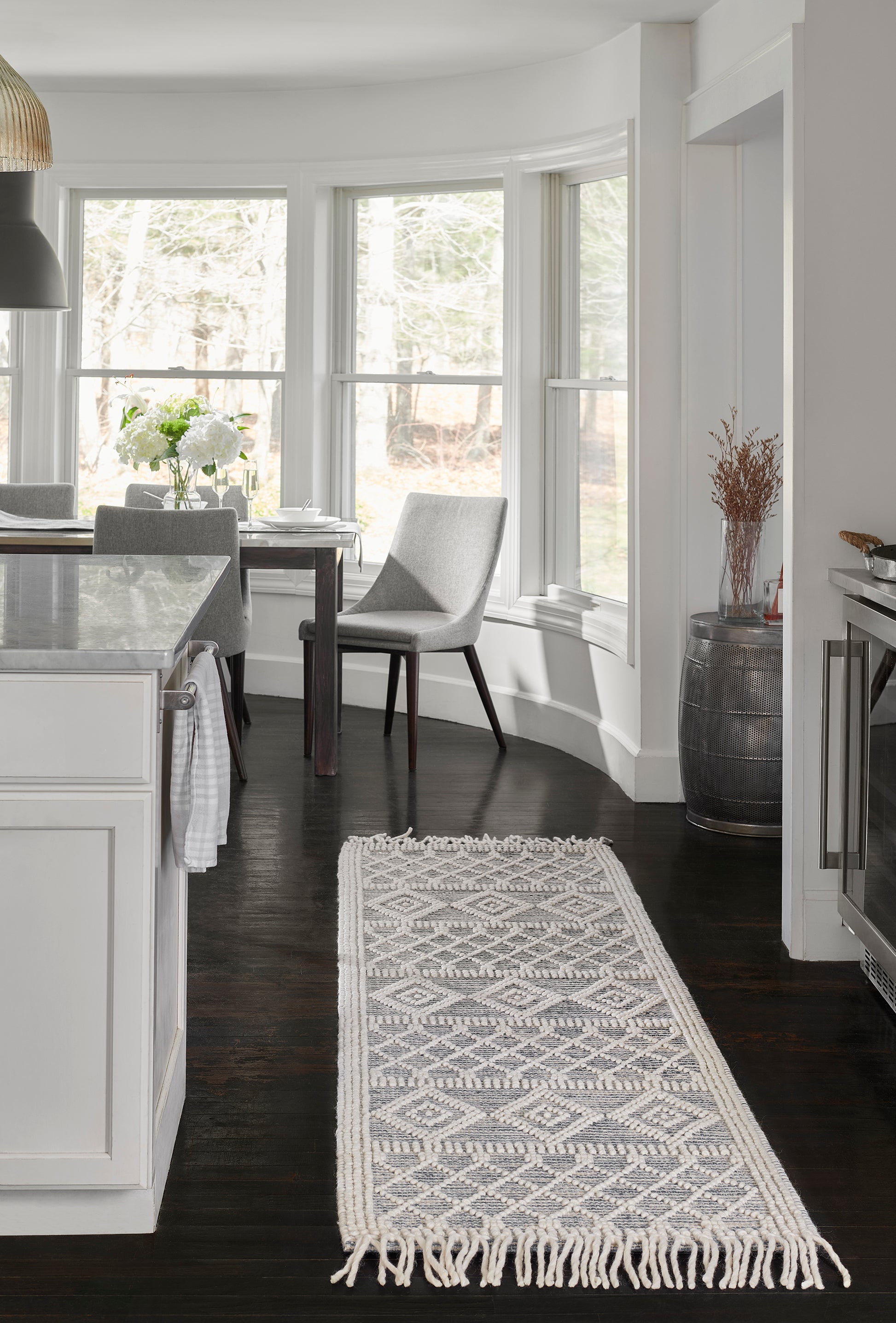 Grey and white woven geometric area rug with fringe on dark hardwood in a bright white kitchen and dining area.