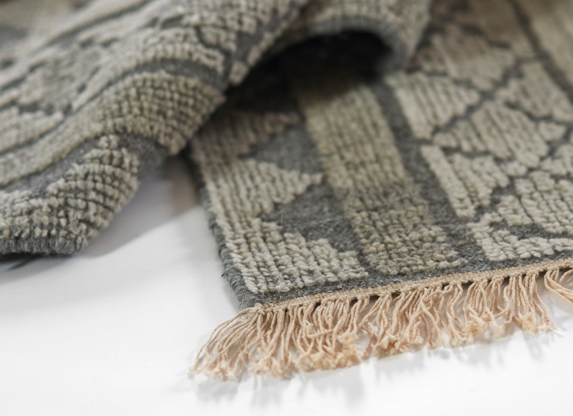 Close-up of gray geometric rug with beige fringe along edge, textured woven pile.