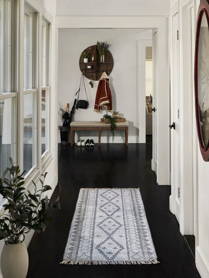 Hallway with dark wood floors and a gray geometric rug, bench with coat rack, circular shelf, plants, and shoes.