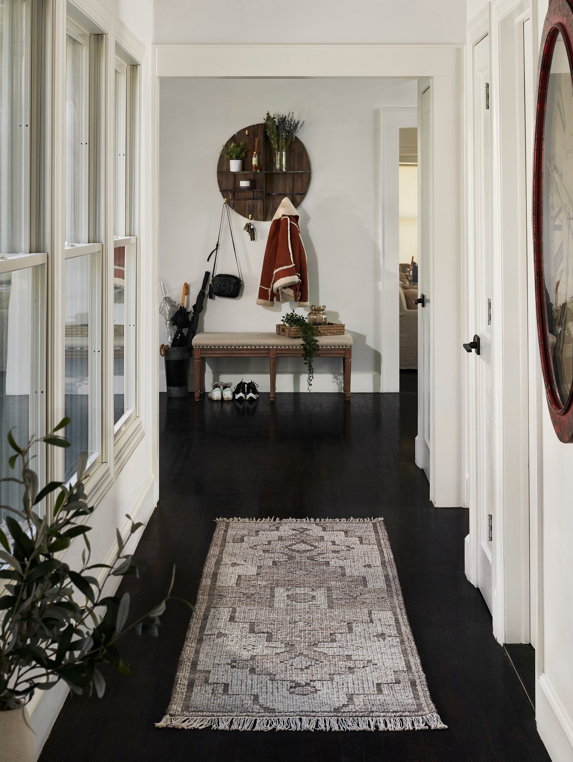 Narrow hallway with dark hardwood, beige-gray geometric runner rug with fringe, white walls, bench and round wooden shelf at end.