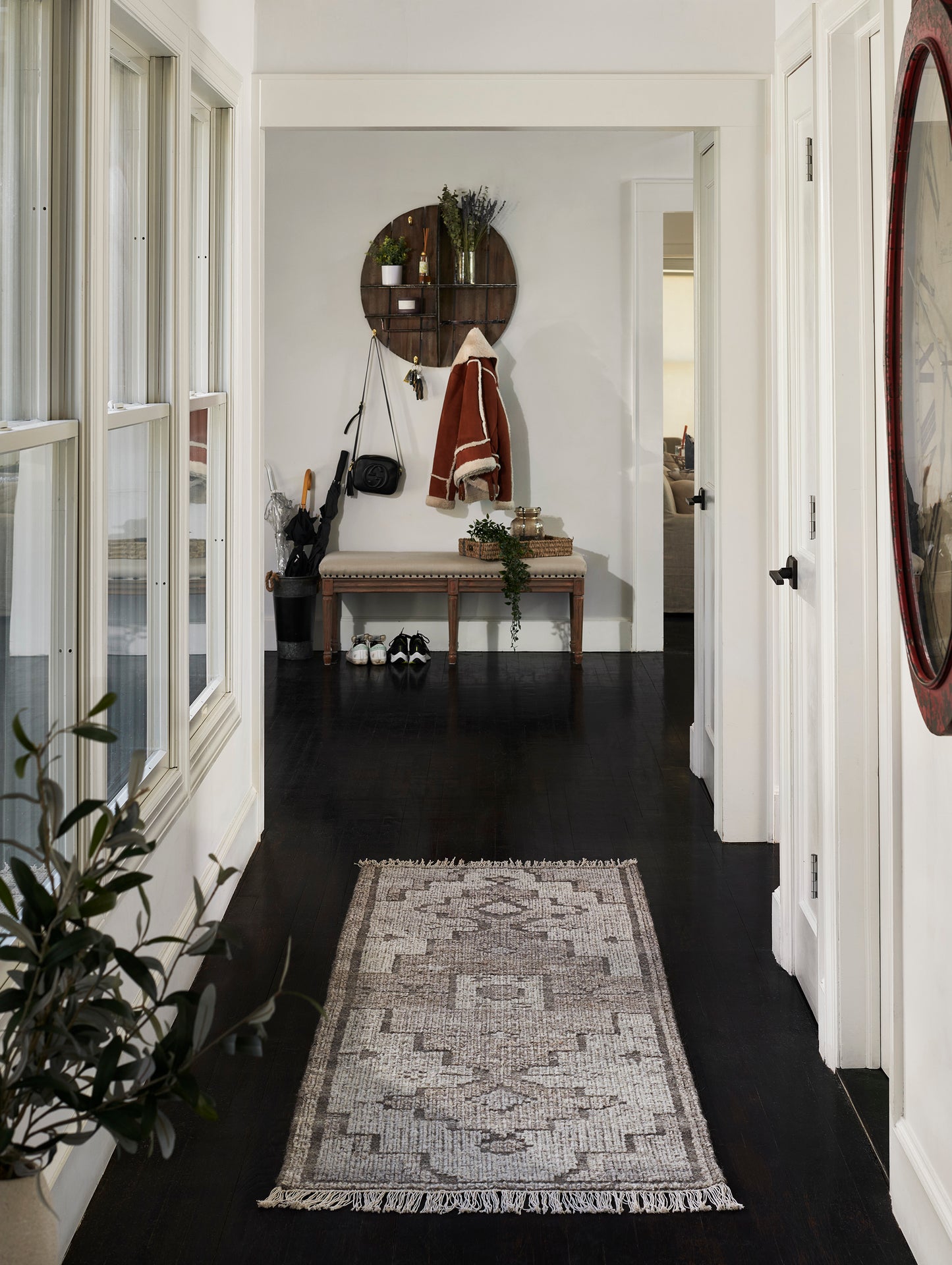 Hallway with dark hardwood floor and white walls; pale patterned runner rug, wooden bench, circular wall shelf with coats.