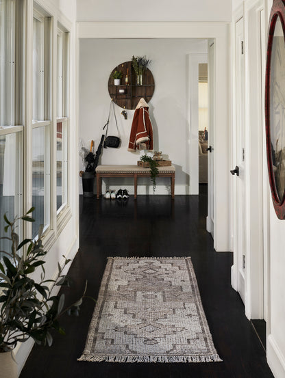 Hallway with dark wood floor, beige geometric rug, white walls; bench under round wooden shelf with coat; red-framed mirror.