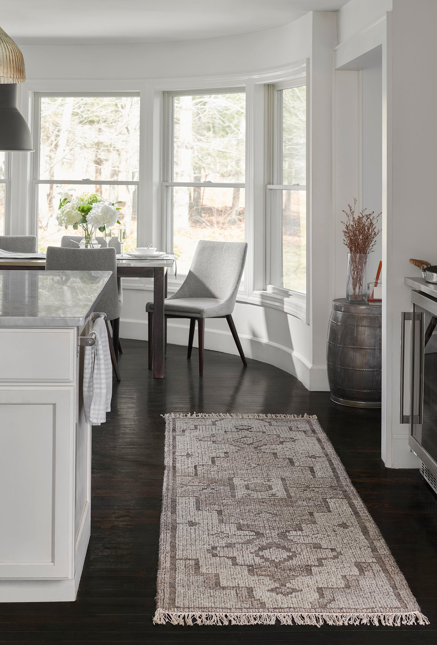 Neutral geometric rug runner in beige and gray with fringe, on dark hardwood beside white kitchen island and bay-window dining area.