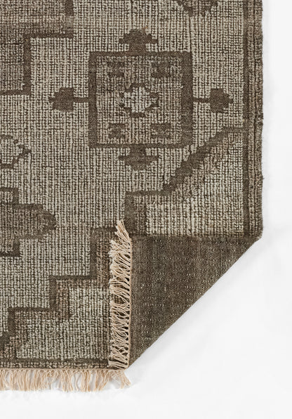 Brown geometric area rug with woven texture, beige motifs, and fringed edges; corner folded showing reverse side.