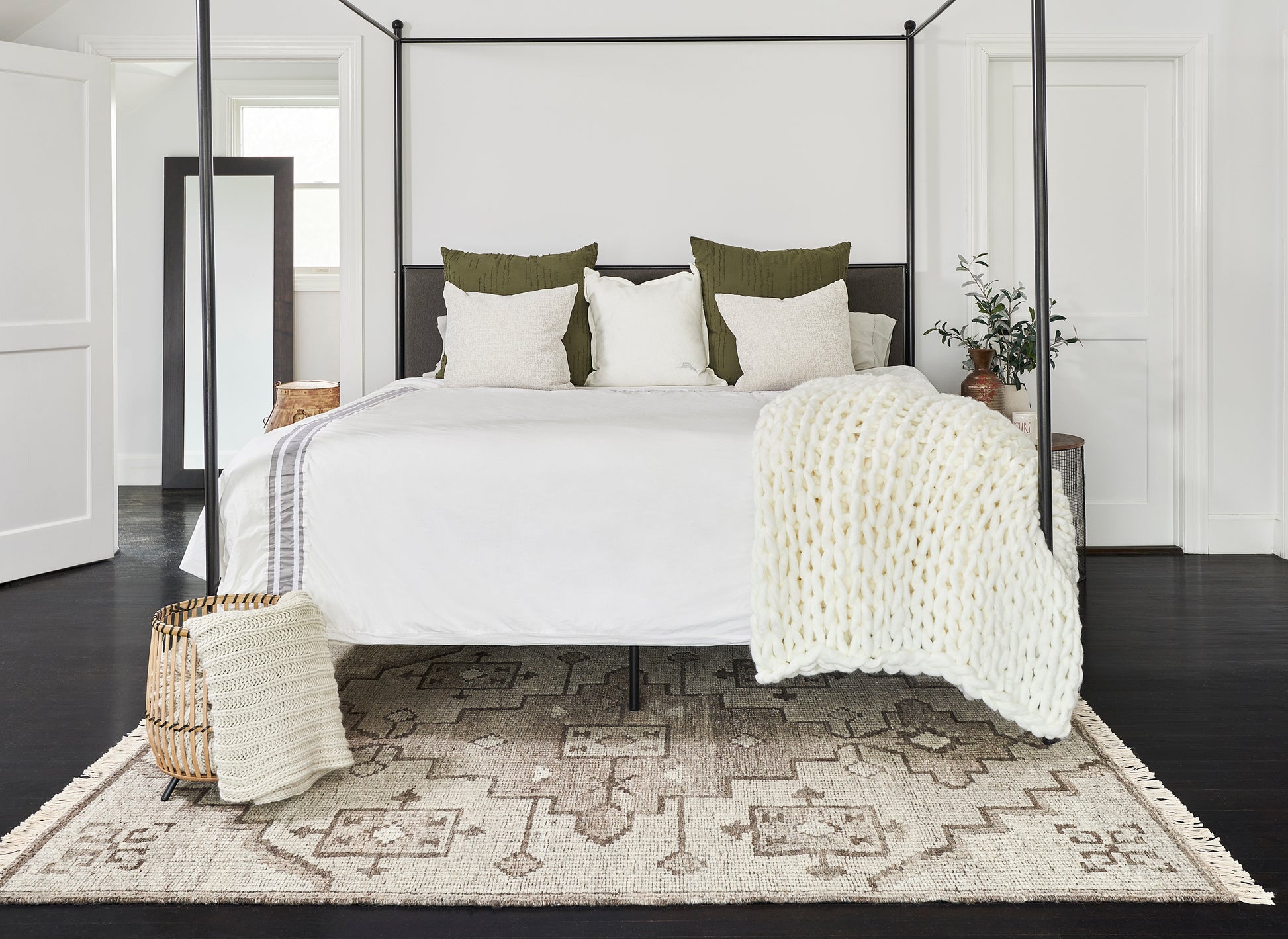 Black metal four-poster bed with white bedding, olive cushions, chunky white knit throw; woven basket & pouf, beige rug.