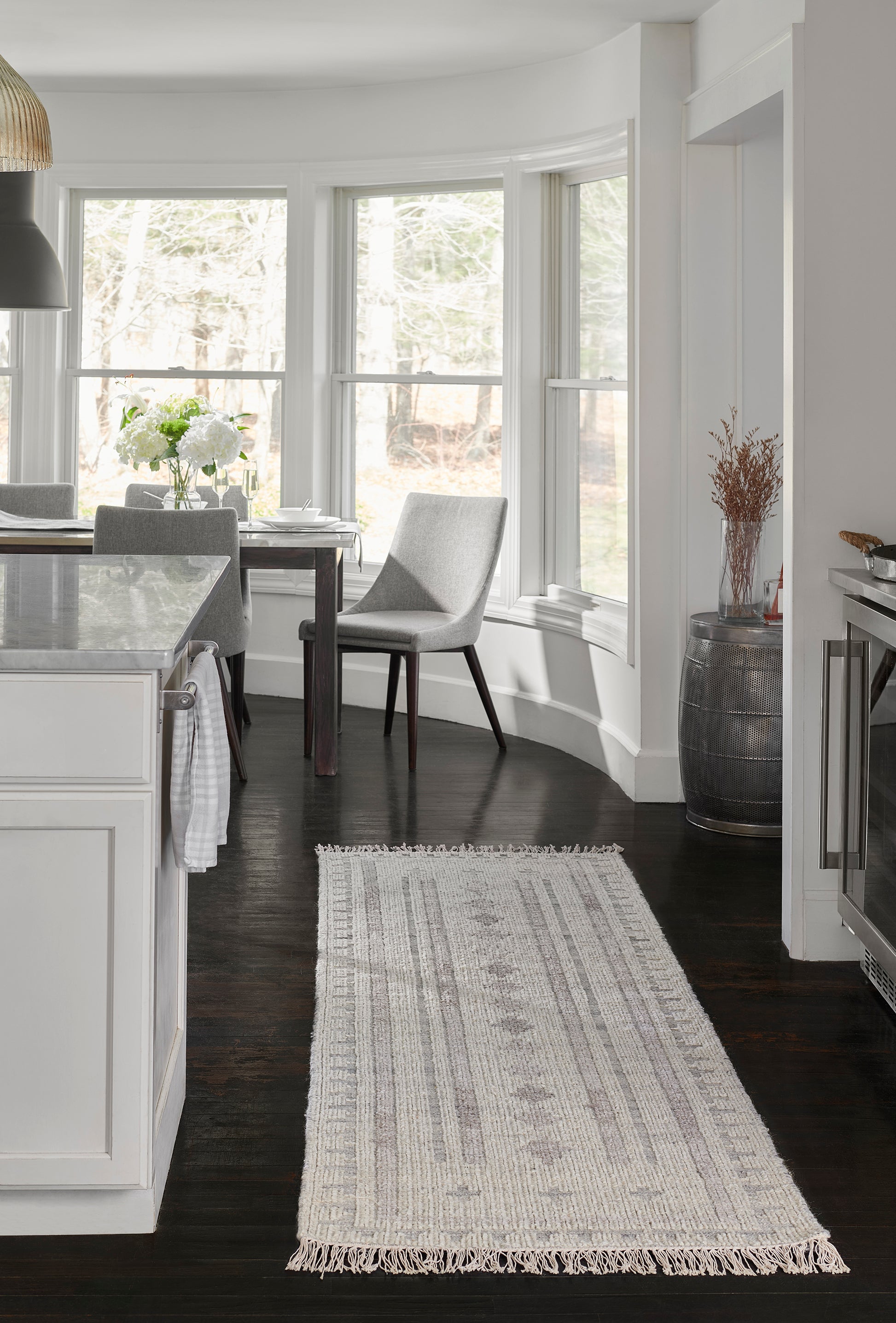 Long woven cream-gray rug with fringe over dark hardwood in a white kitchen with marble island and gray chairs.