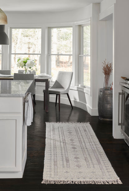 Long woven cream-gray rug with fringe over dark hardwood in a white kitchen with marble island and gray chairs.