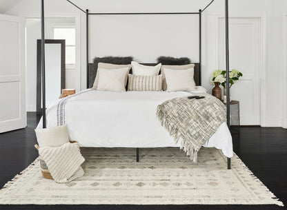 Black metal four-poster bed with white linens, beige pillows, textured throw, cream rug, and vase of white flowers.